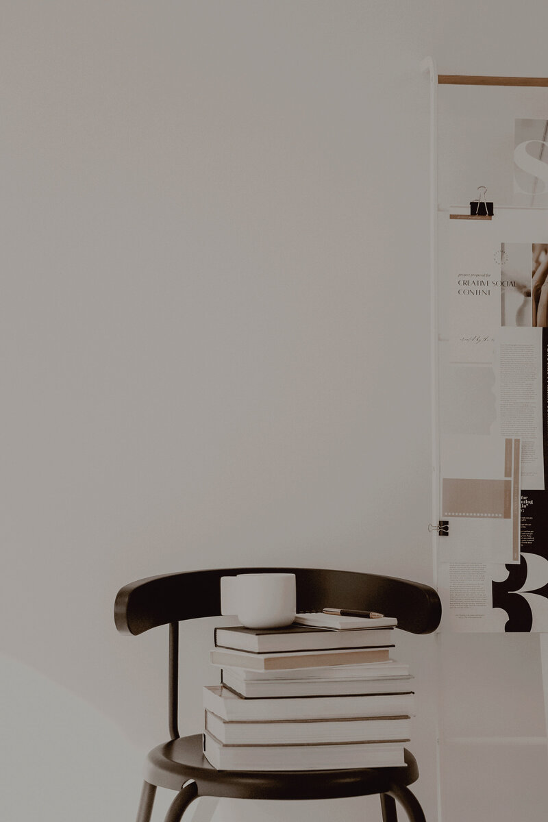 Minimalist workspace featuring a black chair stacked with books, a white coffee cup, and a gold pen. A mood board with creative content inspiration is clipped to a white stand in the background—capturing Sable & Rose’s focus on thoughtful PR strategy, creativity, and intentional branding.