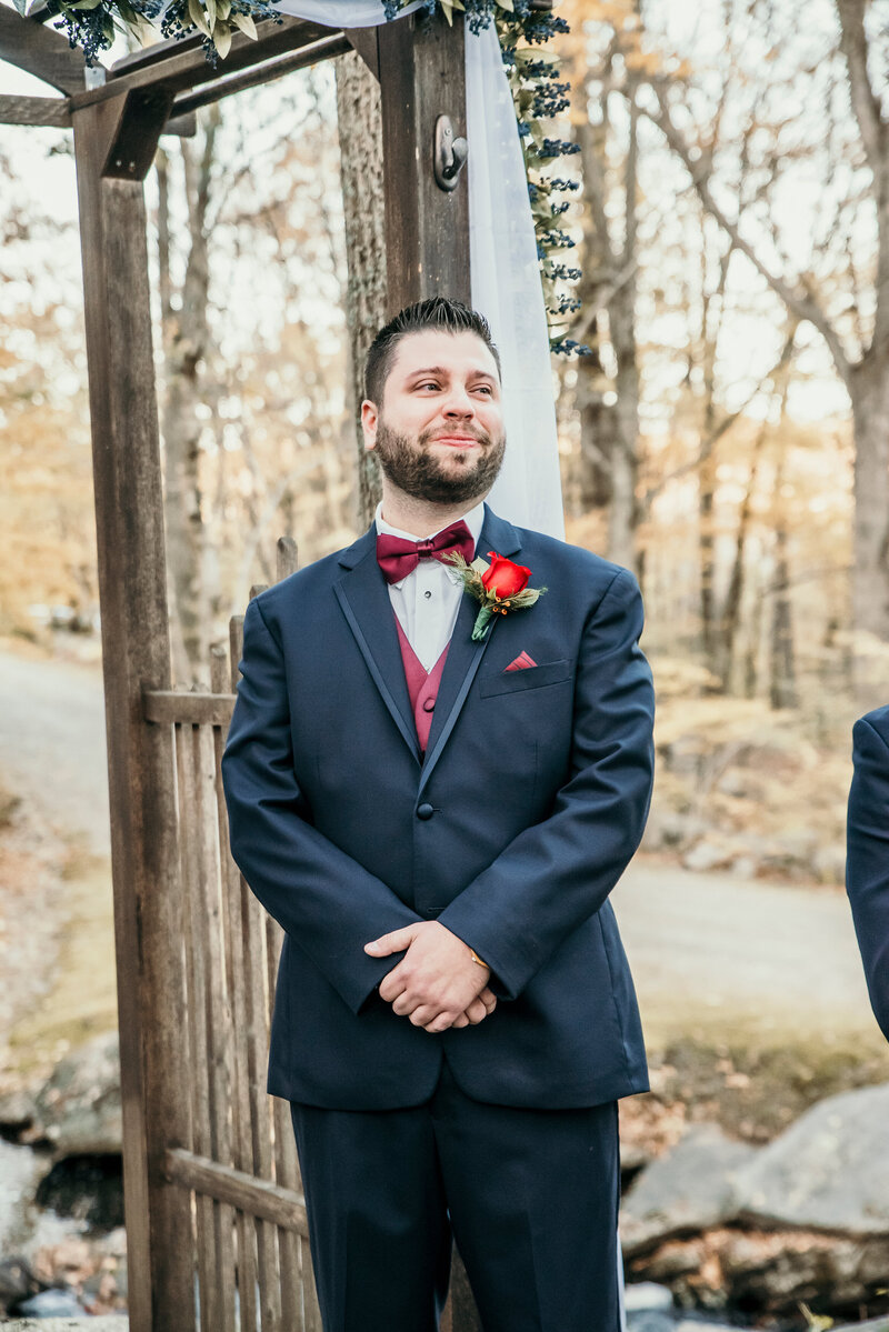A groom in a navy suit and burgundy bow tie smiles with anticipation as he waits at the altar during an outdoor wedding ceremony in Connecticut. Captured by Nicole Mele Photography, documenting the emotions of the big day.