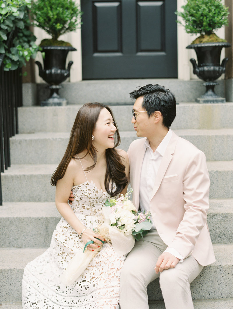 A bride and groom smile and laugh while sitting on a stoop.