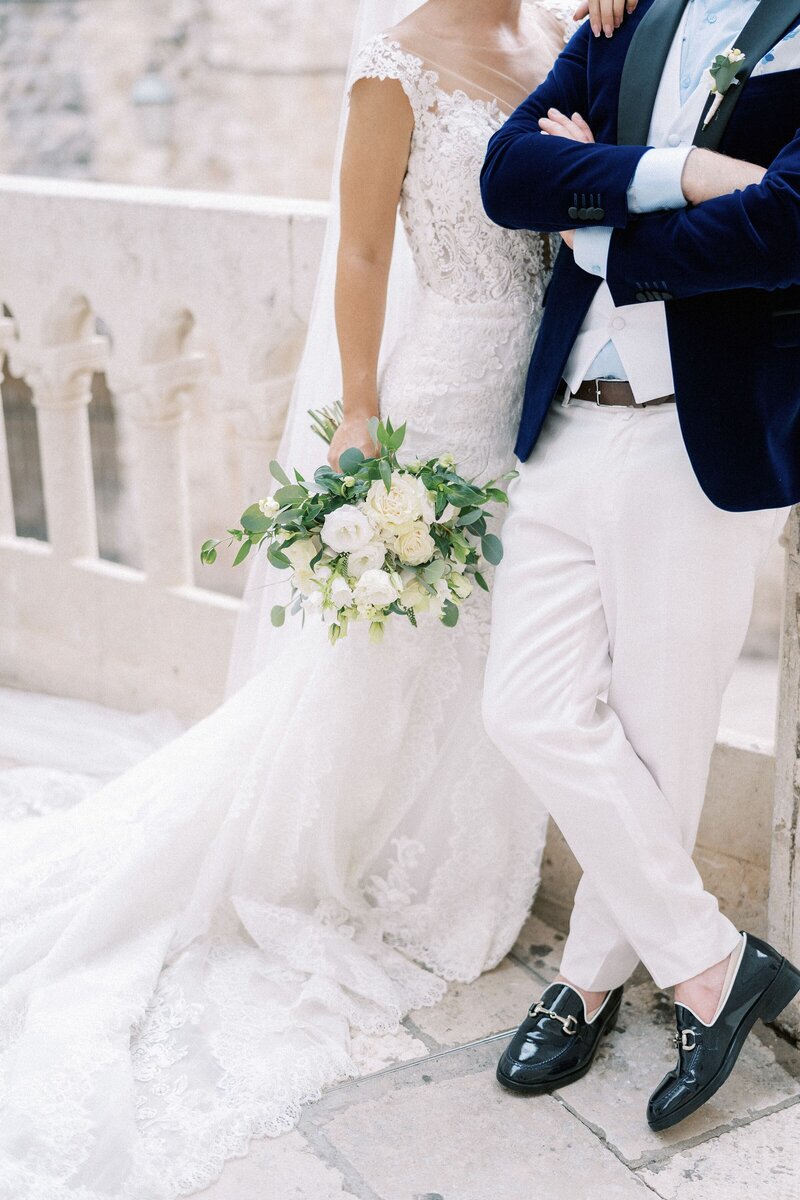 bride-with-bouquet-stands-with-her-hand-shoulder-groom-who-has-his-arms-crossed-his