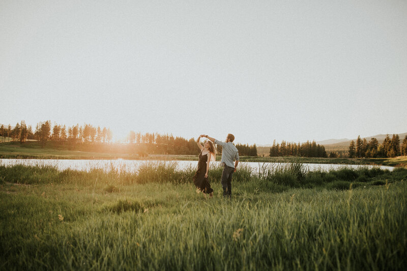 A man twirls a woman around in the grass in front of a lake