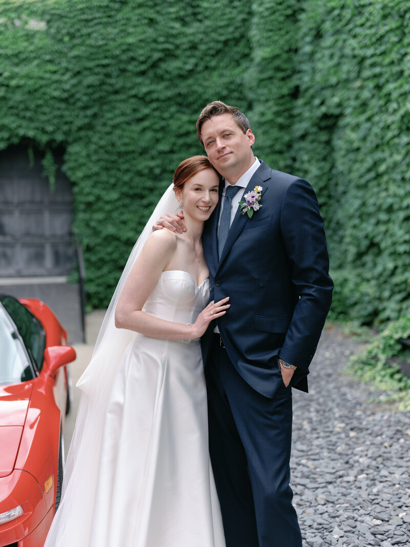 Bride and groom smile at the camera outdoors next to red car.