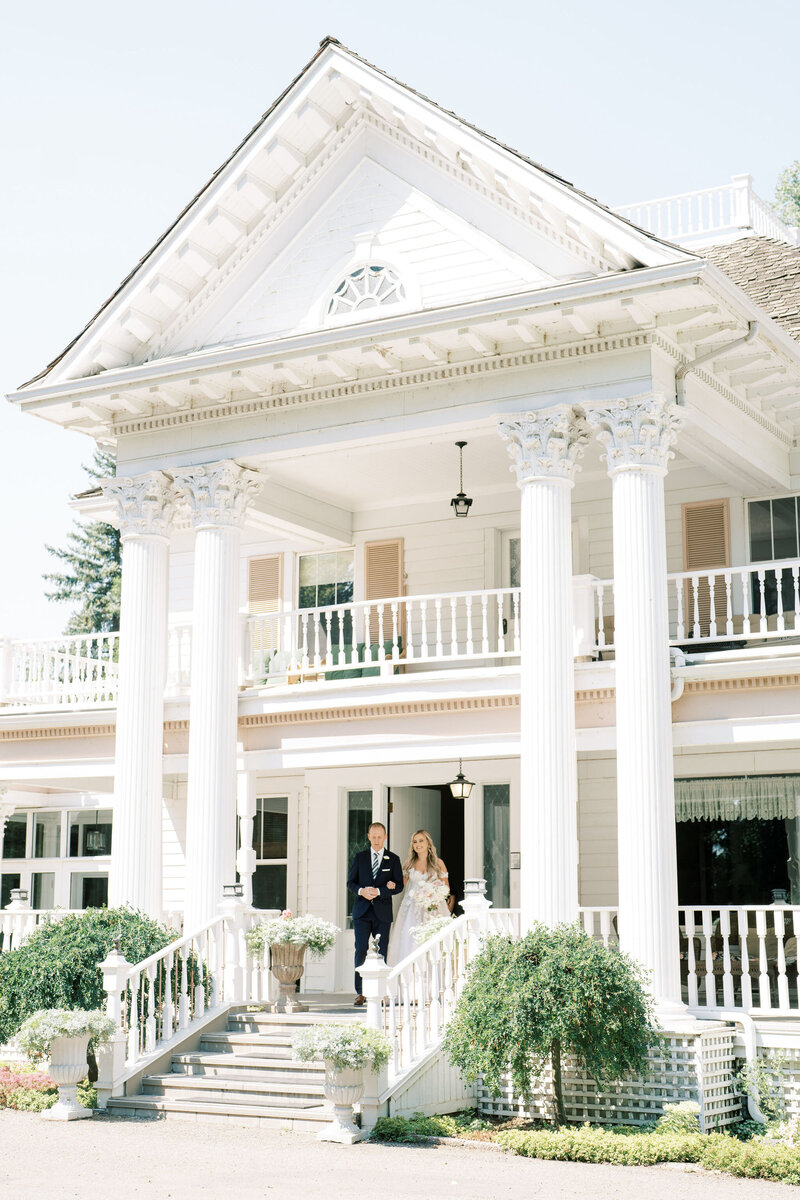 A bride and groom descend the steps of a large, white, columned building on a sunny day, the perfect culmination of their journey with exceptional wedding planning services.