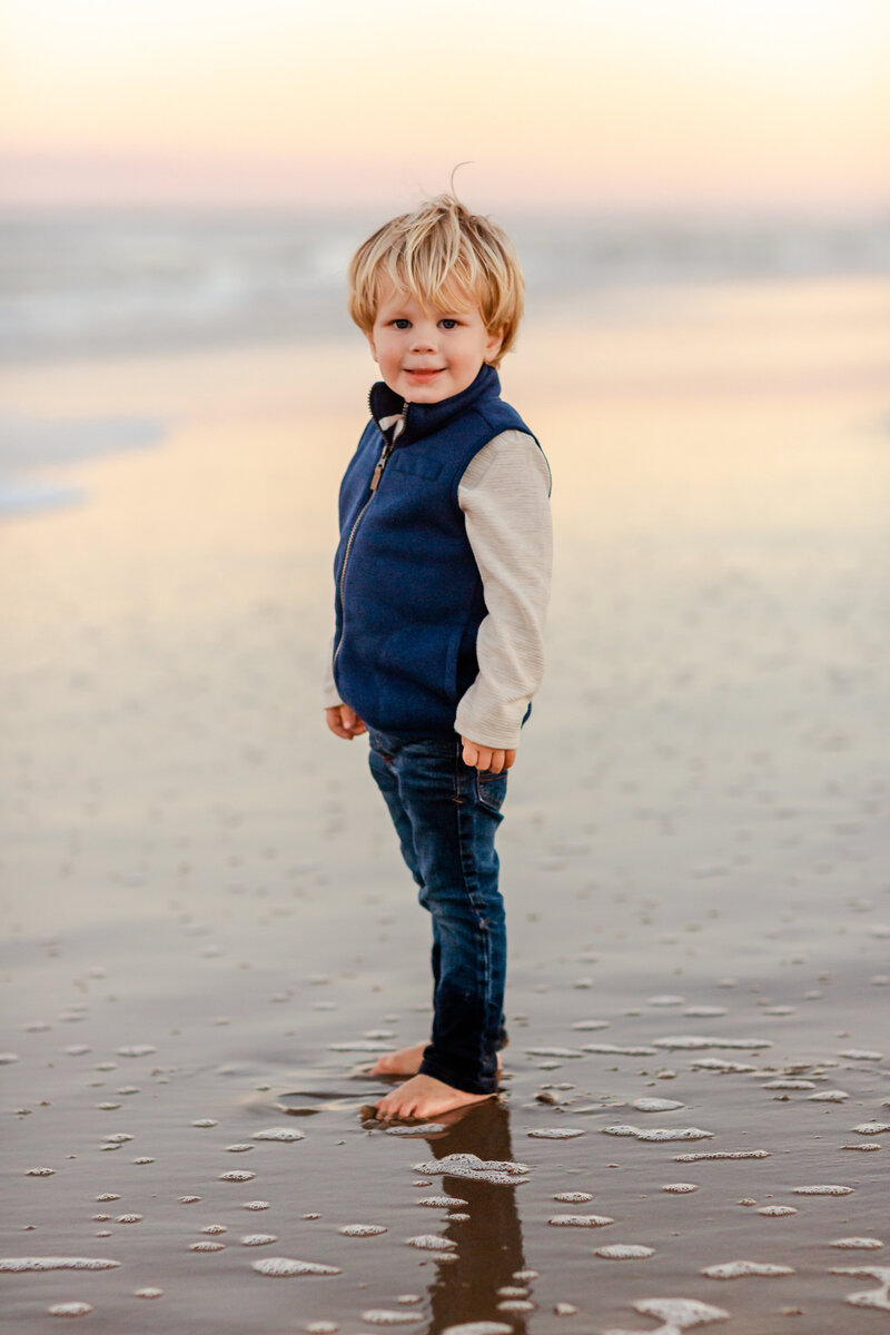 A young boy stands barefoot at the shoreline, pausing to watch the waves during a quiet moment captured by Brooke Tucker at sunset on the Virginia Beach Coast