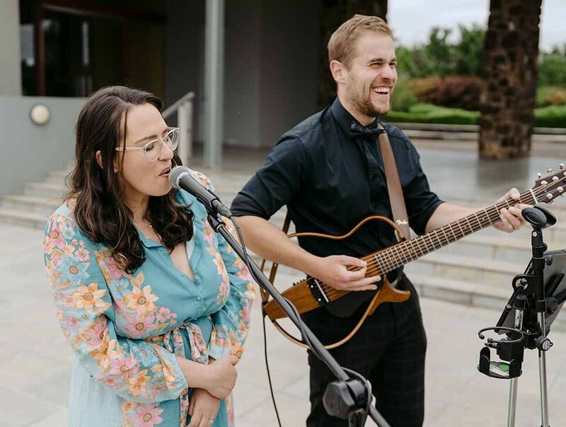Female singer and male acoustic guitarist performing outdoors, casually dressed, creating a relaxed and joyful vibe.