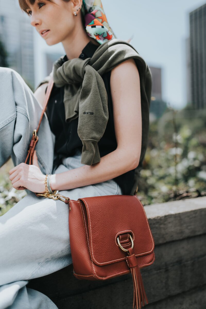 Woman with colored scarf in her and bangs wears a brown purse and a green sweater tied over her shoulders as she sits outside and looks off-camera
