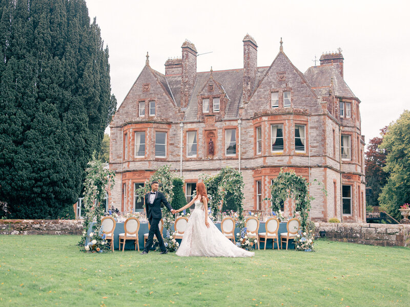 Bride and groom walking across lawn holding hands with their wedding reception table and Castle Leslie Estate behind them.