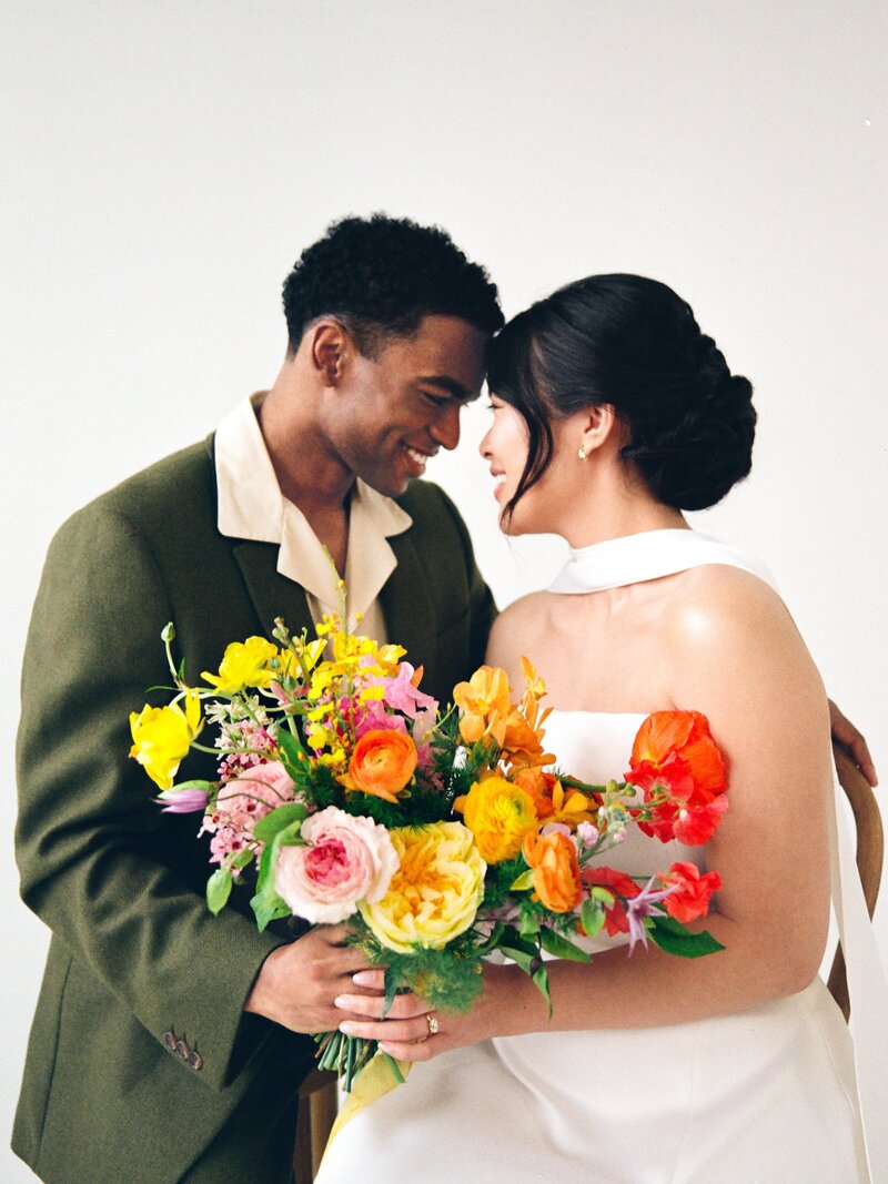 Smiling couple embracing, holding a vibrant, textured bouquet with poppies, roses, and other seasonal blooms in orange, yellow, and pink hues.