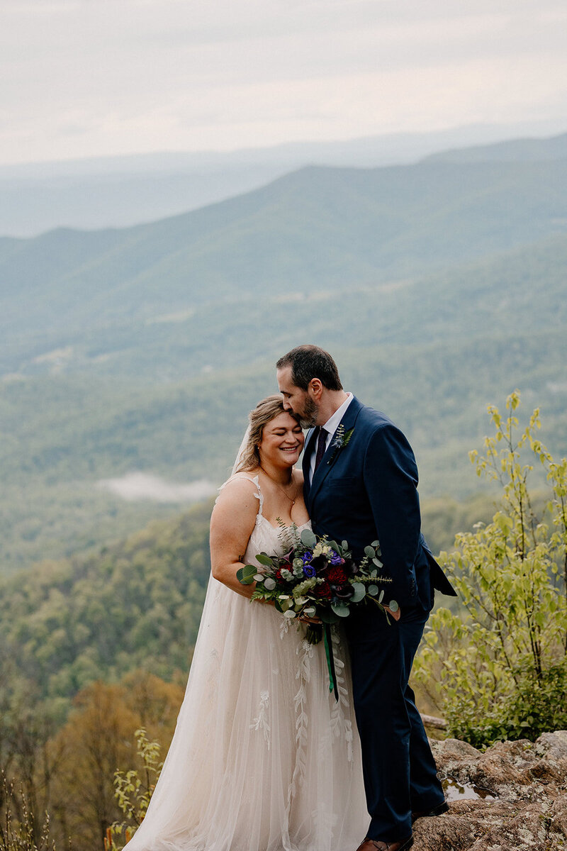 A couple stands in front of the Blueridge Mountains on their elopement day. The groom is kissing the forehead of his bride. 