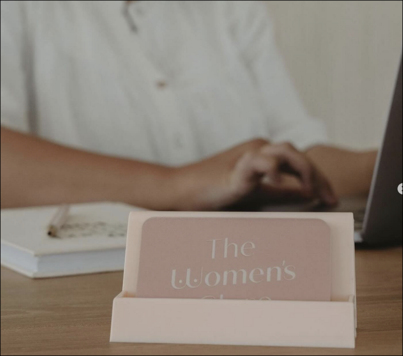 a woman at a desk typing on a laptop. in the foreground is a stand with business cards which are a pale pink with the words the womens