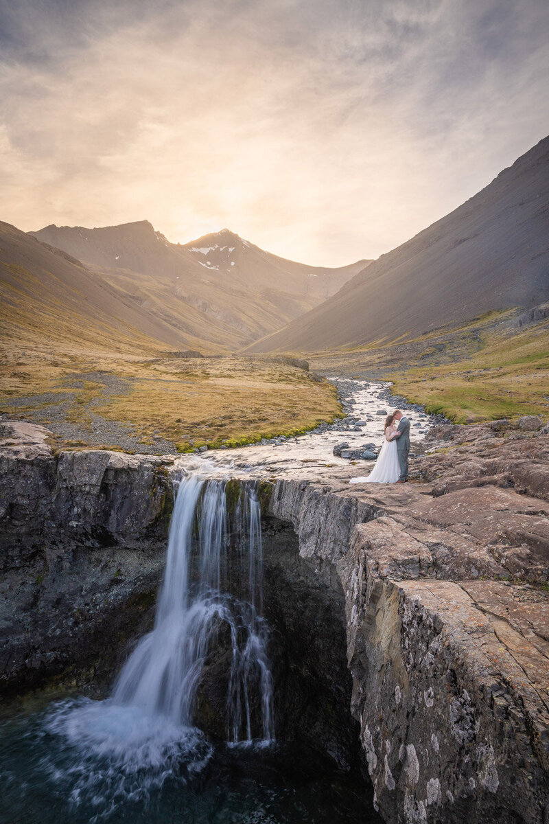 A couple in their wedding attire standing next to waterfall in Southeast Iceland at sunset. The valley is surrounded by mountains. 