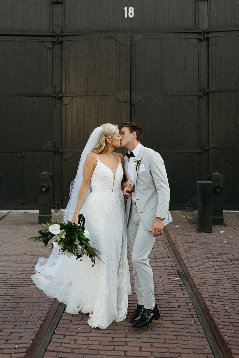 A bride in a white gown and a groom in a light gray suit share a kiss, beautifully captured by an Ontario destination wedding photographer at Steam Whistle Brewing in Toronto. They stand on brick pavement with visible train tracks, framed by large industrial doors—a perfect moment for timeless wedding film photography.