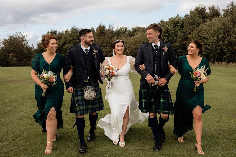A bride and groom are photographed waking with their wedding party in a green Aberdeenshire field. They link arms and smile.