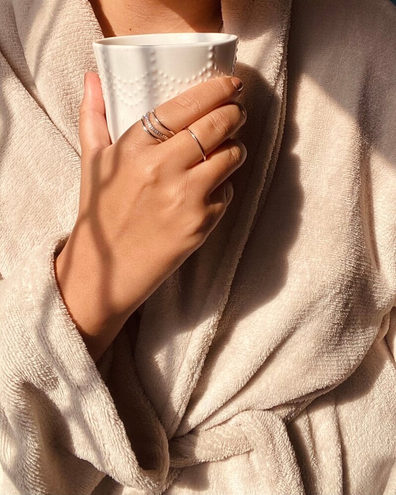 A person in a cozy beige robe holds a textured white mug with sunlight casting soft shadows. The scene conveys a warm, relaxed morning atmosphere.