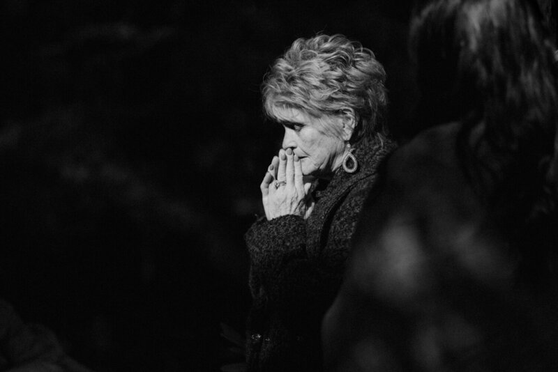 Black and white photo of a wedding guest pausing with hands clasped in emotion during the ceremony, captured in a timeless documentary style by Rainstorm Photo & Video.