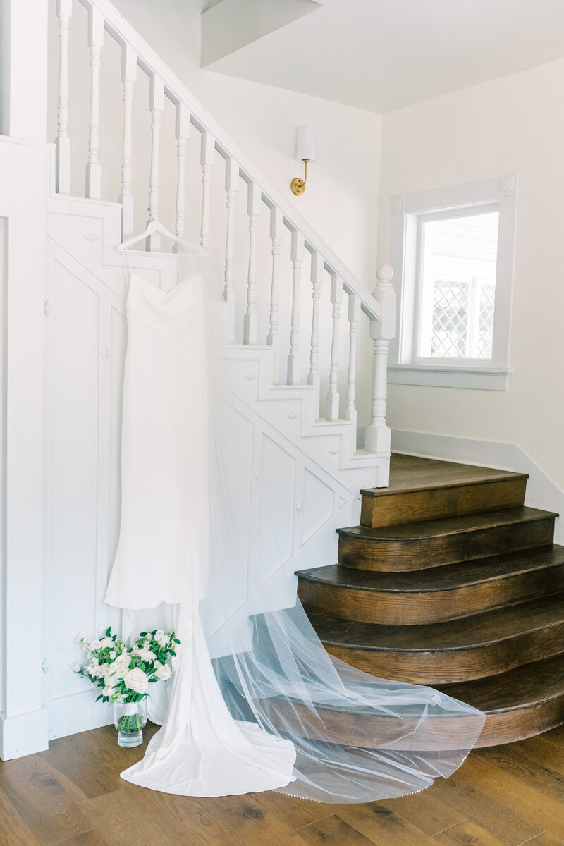 Wedding dress hanging on a staircase railing with the veil draped on the steps and the bouquet in a vase on the floor