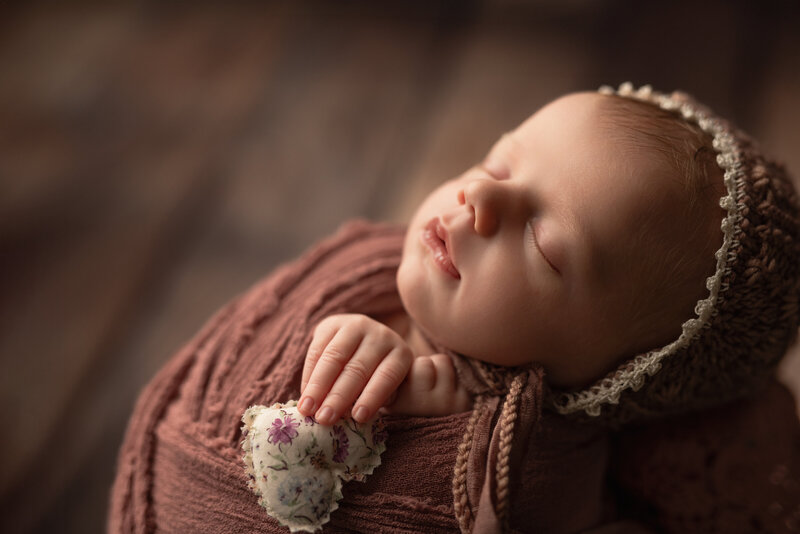This heartwarming close-up captures every tender detail of a sleepy newborn girl in dusty rose, holding a handcrafted heart.