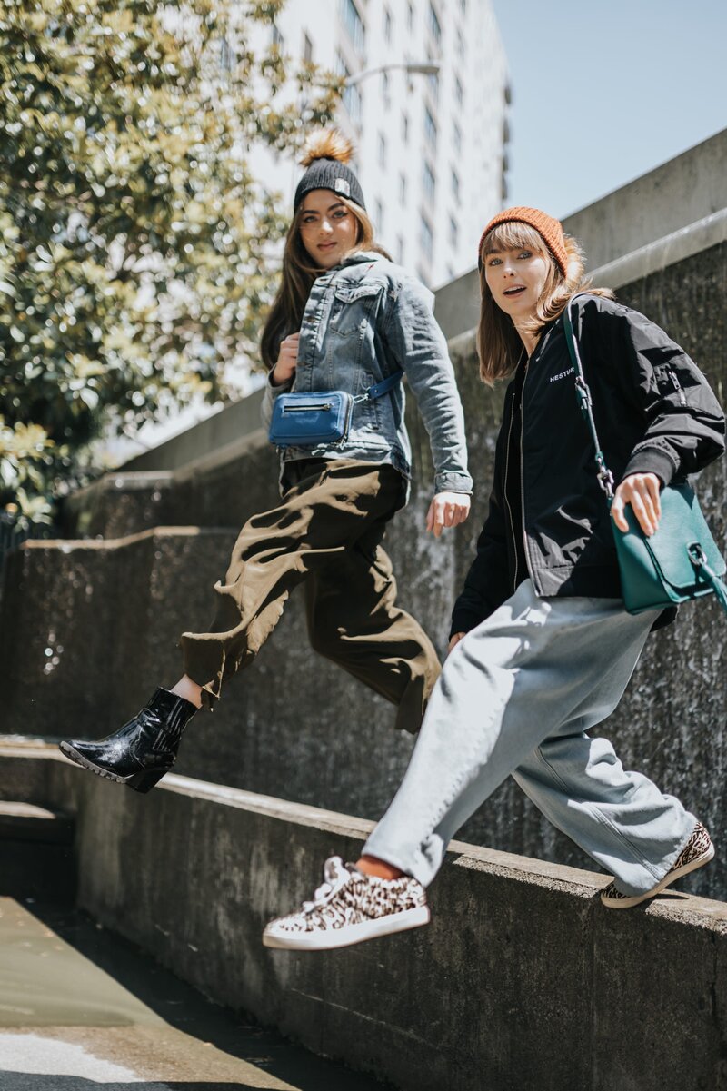 Two women with colorful purses jump mid-air from a short cement wall.