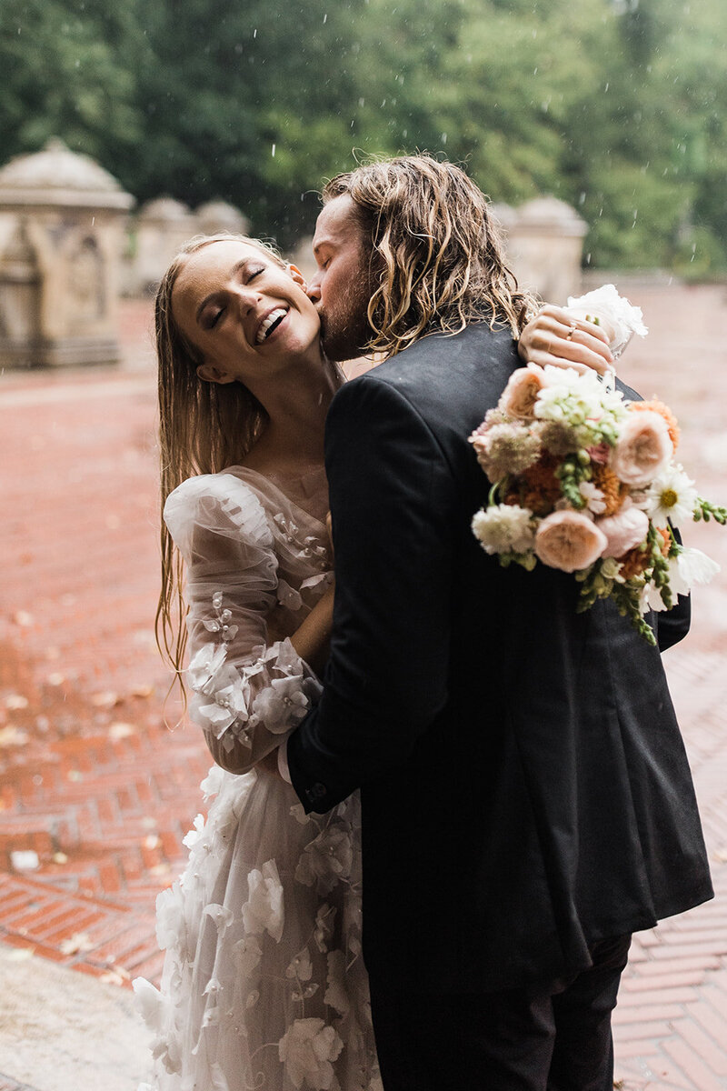 Bride and groom in the rain in central park in new york city, bride is holding her flowers and the groom is kissing her cheek