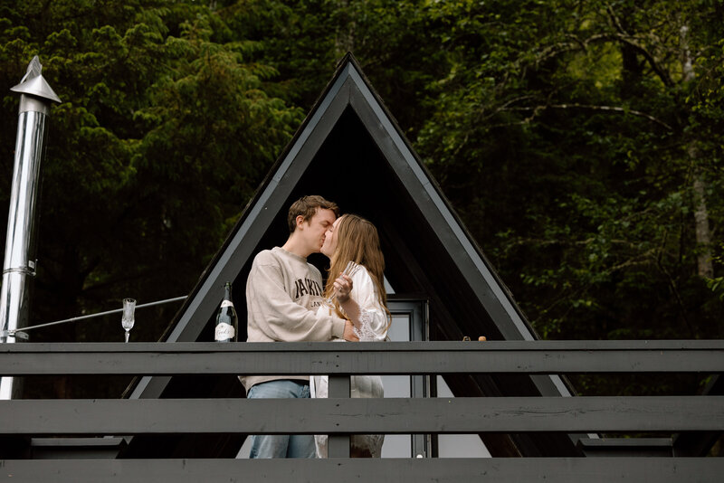 Couple toasting champagne while standing on the deck of their a-frame cabin.