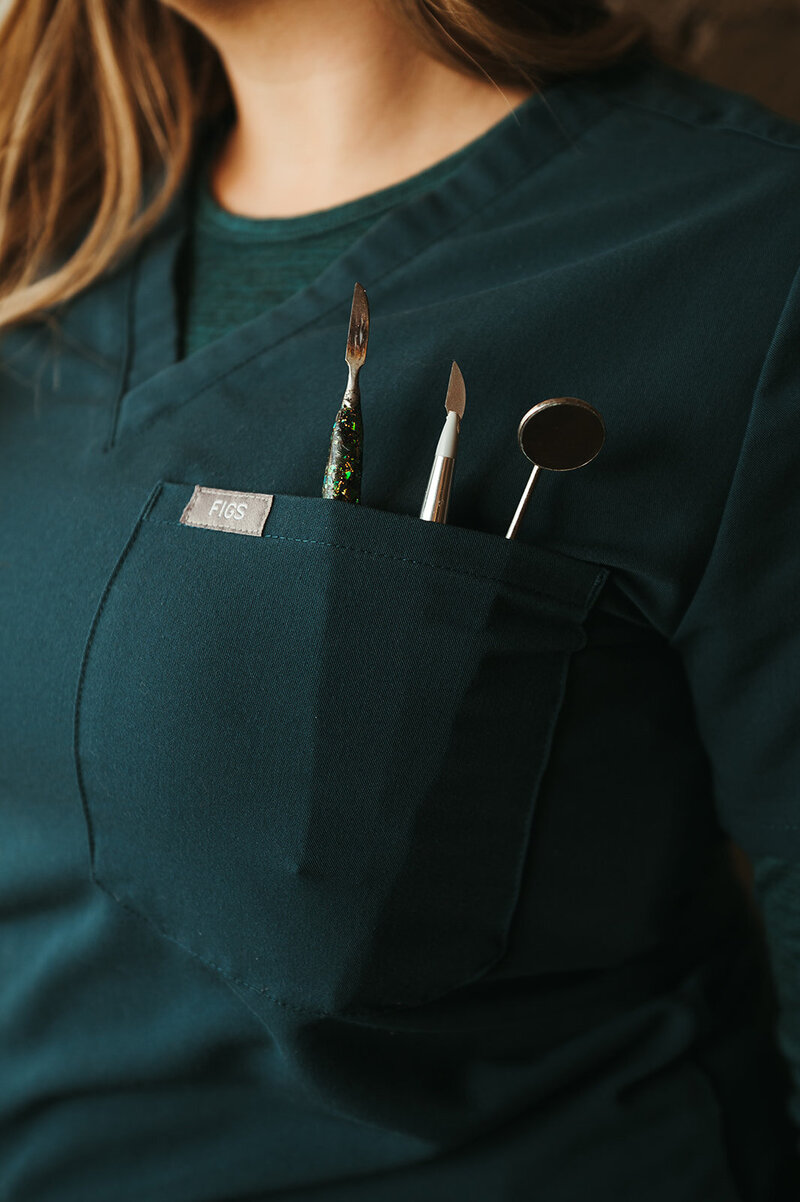 A close up of a woman wearing teal colored scrubs, with denture creation tools tucked in her chest pocket.