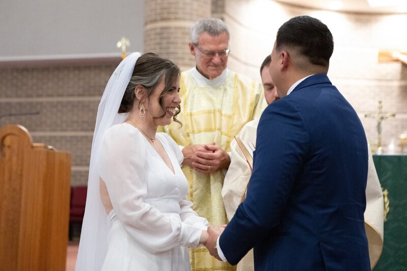 A bride and groom hold hands as they say their vows and priests look on.