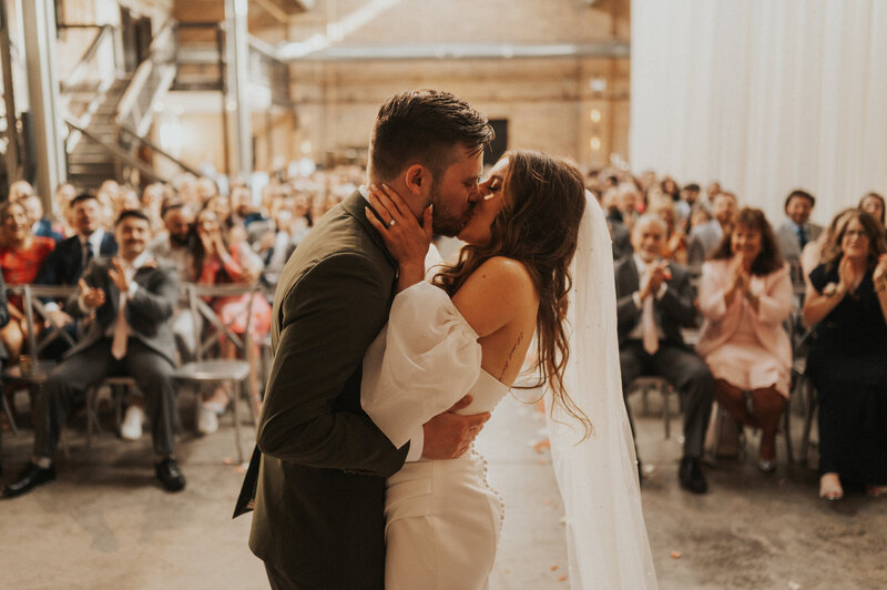 A couple kisses passionately at their indoor wedding ceremony, surrounded by applauding guests seated in rows in Chicago, Illinois.