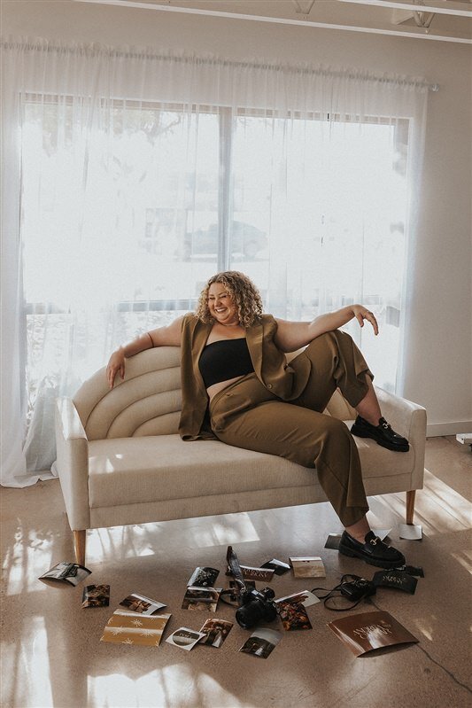 Andrea Cakmar lounges on a modern, cream-colored sofa in a bright room. She is dressed in a tan pantsuit with a black crop top, surrounded by scattered photography prints and a camera on the floor.