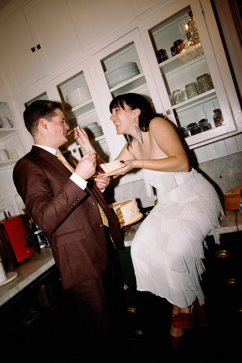 Bride and groom sitting on counter in kitchen sharing cake at their fun and unique Maine wedding