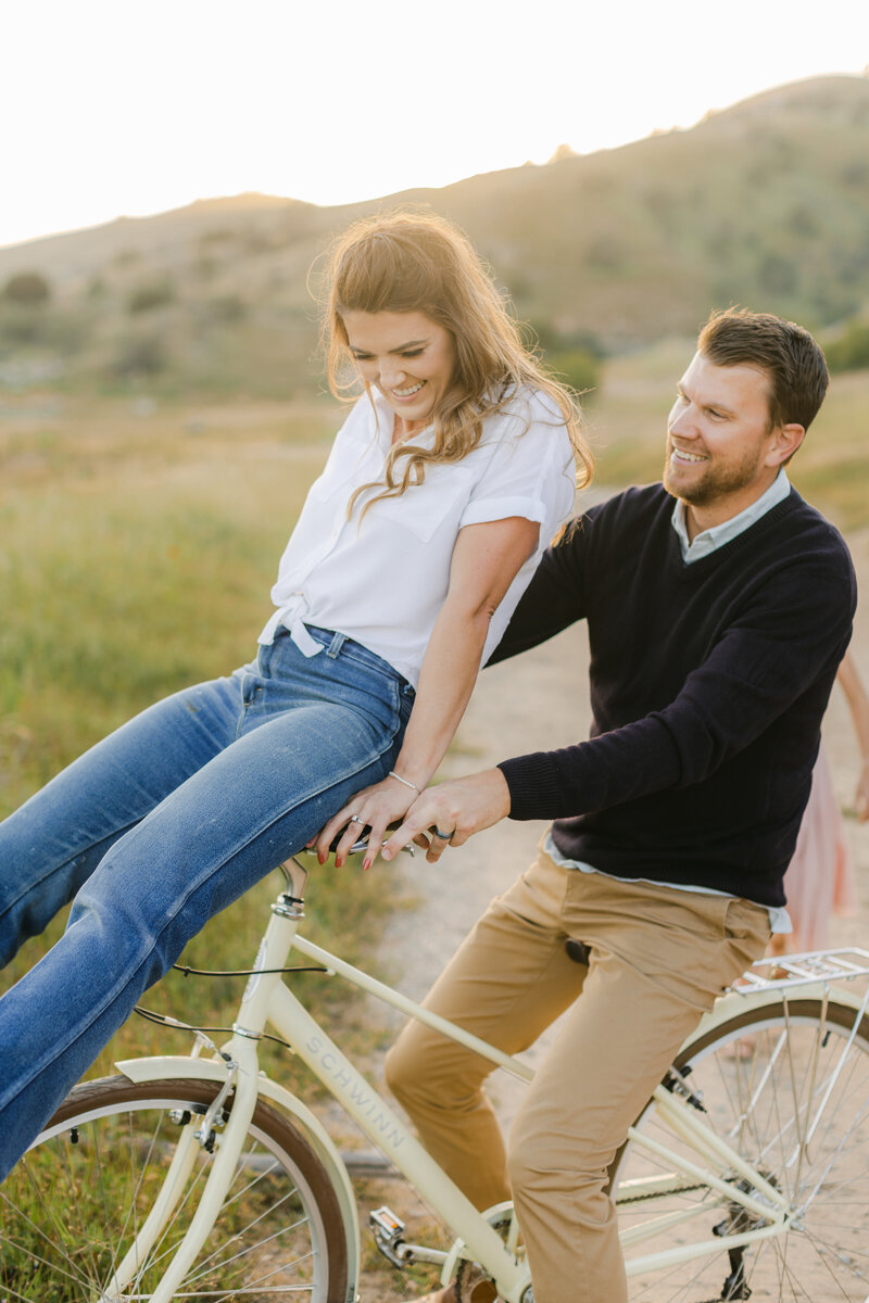 Playful engagement session featuring a stylish couple on a bike ride — she’s laughing on the handlebars with flowy hair and a casual outfit, captured mid-motion with an editorial, carefree vibe.