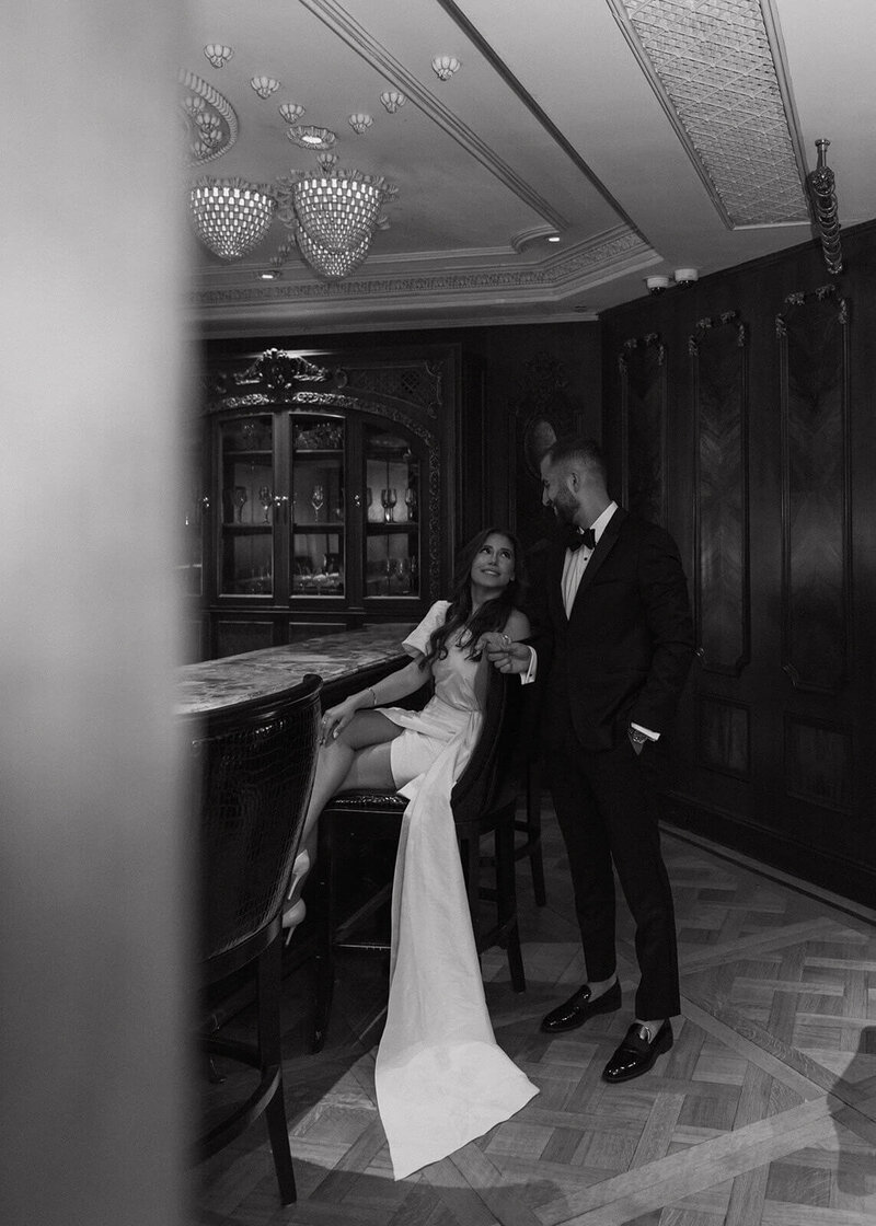 The bride is sitting on a bar stool, and the groom is standing next to her, holding her hand.