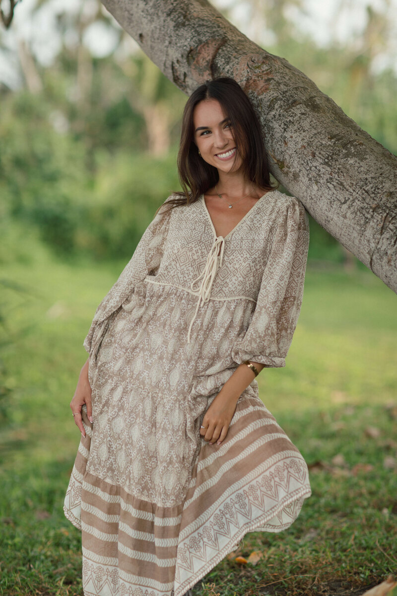 A women smiling and leaning on a tree with a beautiful dress on.