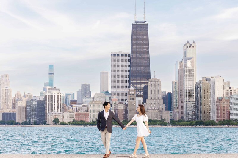 A couple holds hands while walking along the lakefront at North Avenue Beach in Chicago, with the iconic city skyline and John Hancock Center towering in the background on a bright, clear day.