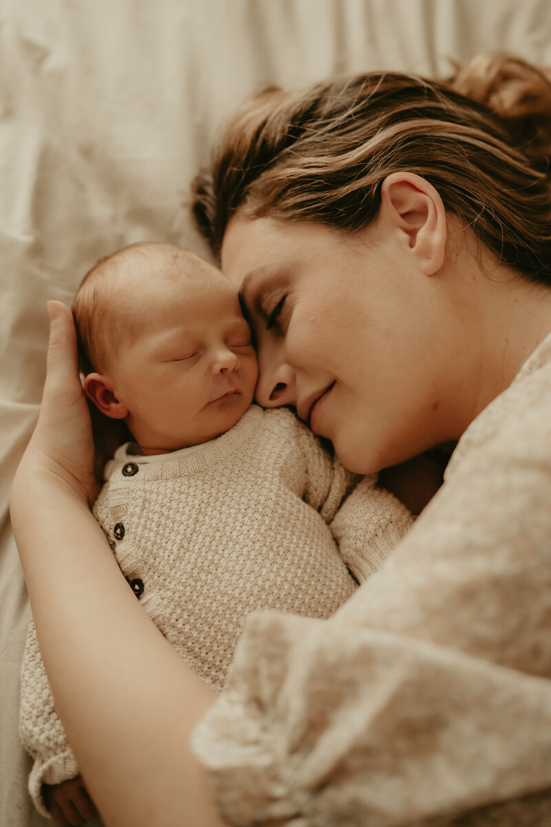 A couple embraces in a golden field during a maternity photoshoot at sunset, with the expectant mother wearing a rust-colored dress and the partner in a white shirt and teal pants, surrounded by tall grass and a scenic natural landscape in Peapack, NJ.
