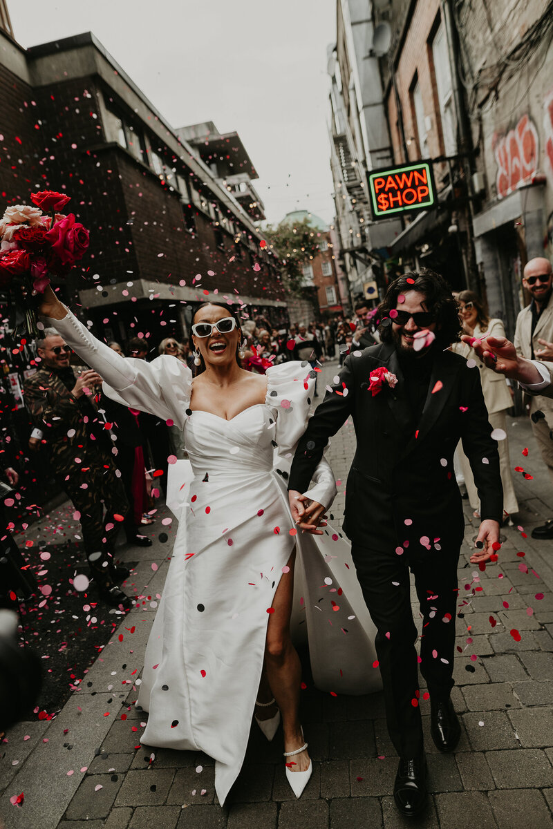 A wedding couple blow pink and red confetti at the camera.