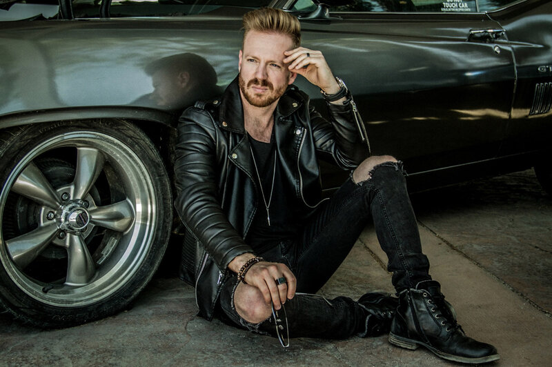 Musician portrait Brad Saunders sitting on ground against classic car