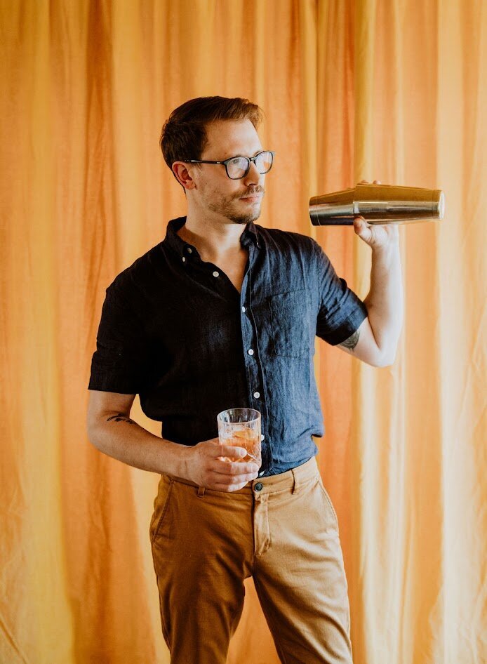 Lee Large holds a shaker and cocktail in his hands ready to shake his next cocktail in front of a yellow velvet backdrop.