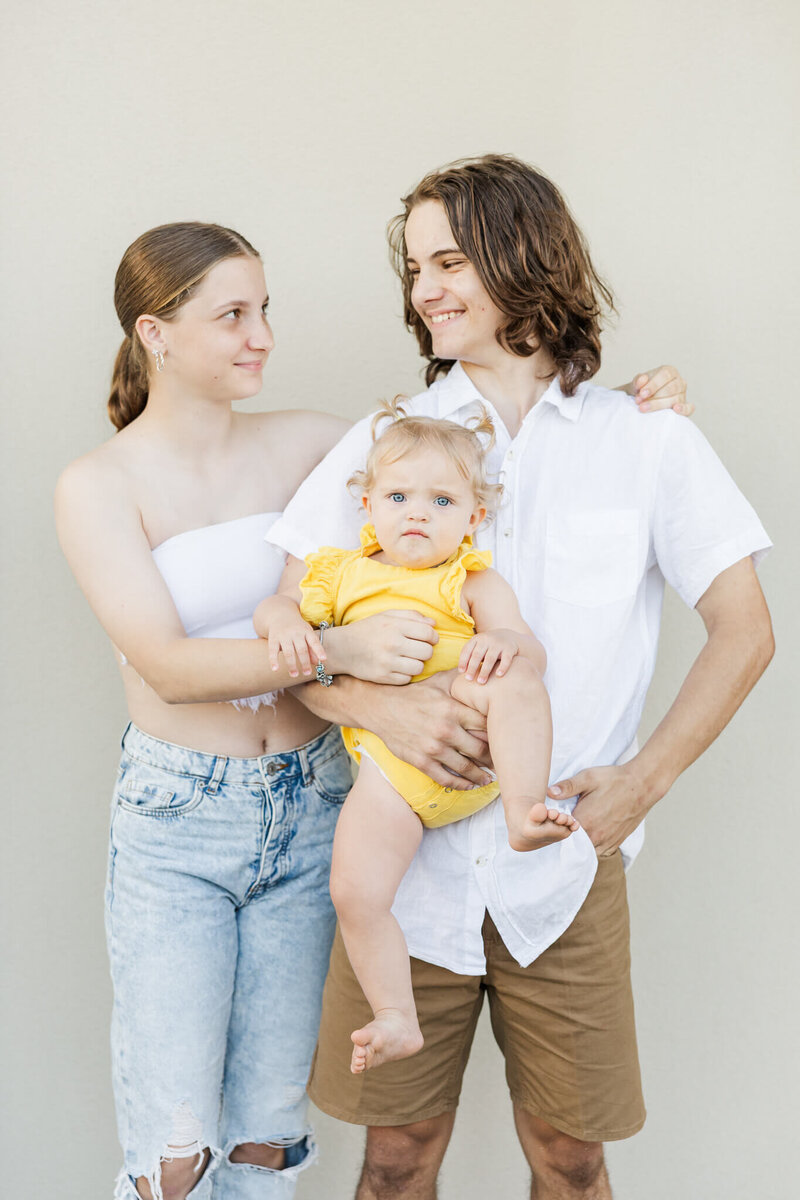Teenage brother with his teenage sister standing next to him while he holds their toddler sister. Located at Rivieria at Cooee Bay Yeppoon.