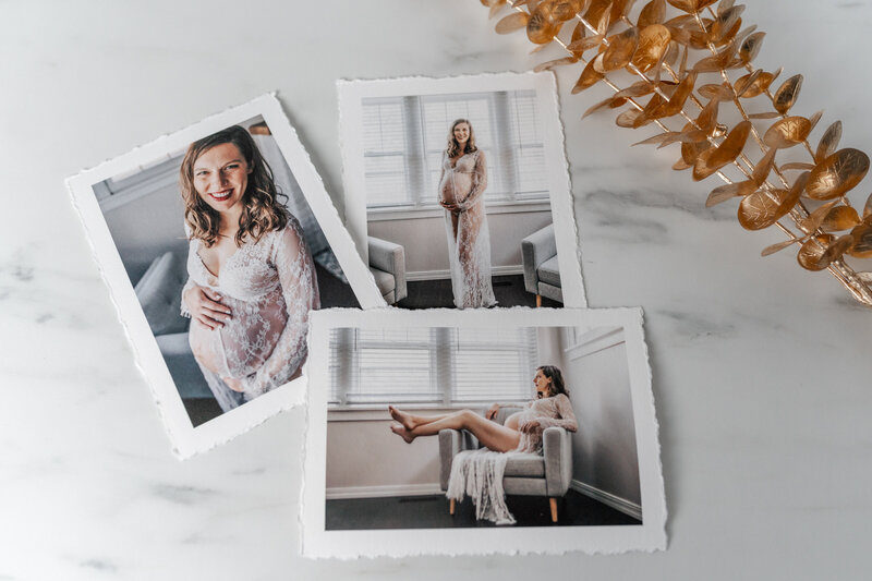 Three printed photographs of a pregnant woman in a lace gown are displayed on a marble surface, with a golden decorative branch placed nearby.