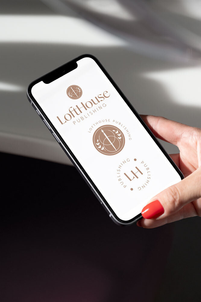 A hand with red nail polish holds a smartphone displaying the logo of LoftHouse Publishing. The design features elegant, modern typography and circular emblems.