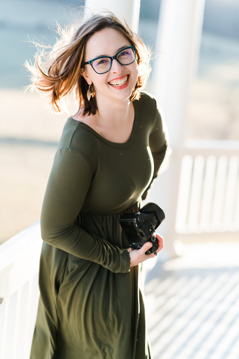 Lively, creative headshot of a photographer posing outside on the front porch, with the sun illuminating her hair and the windy blowing. She is laughing and holding  her camera.