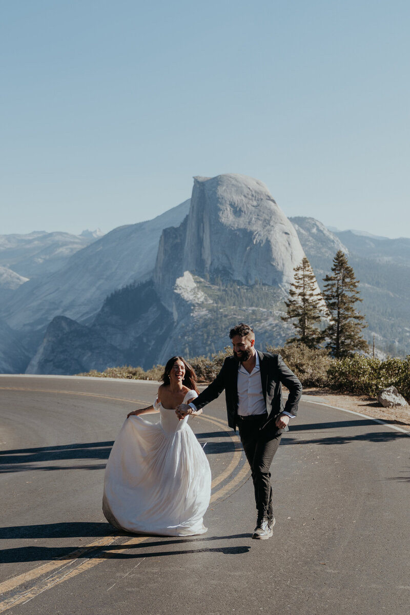 bride and groom running hand in hand down road with half dome in the background during their Yosemite elopement