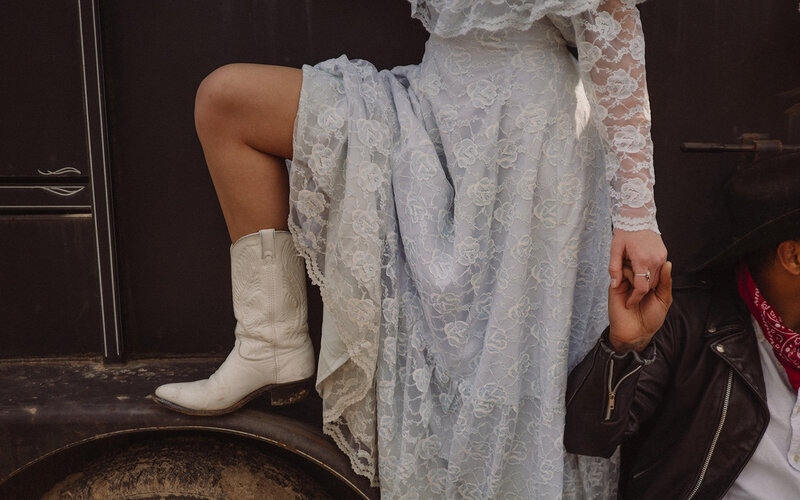 A bride in a vintage blue wedding dress and white cowboy boots holds the hand of a man in a black leather jacket and cowboy hat.