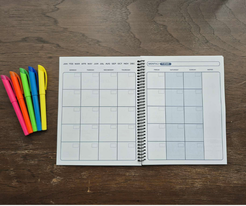 The Time Magic Planner open to the undated monthly spread on a wooden table, with bright highlighters in pink, orange, green, blue, and yellow arranged beside it.