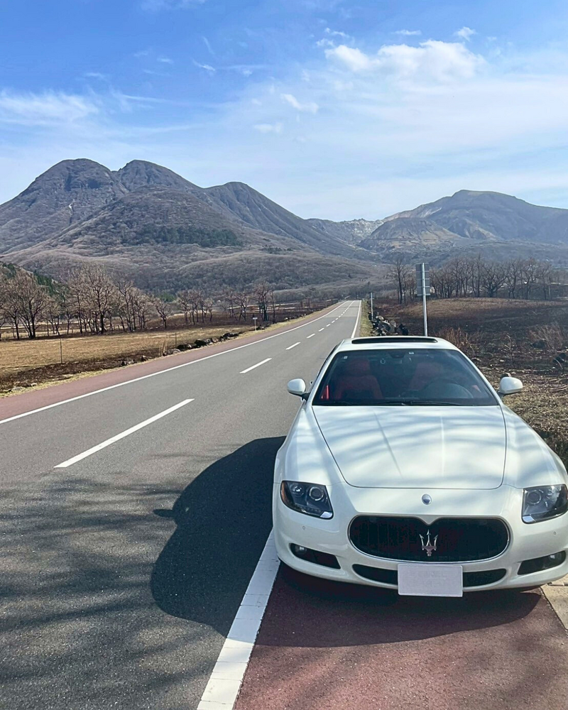 Rear view of a white Maserati driving along an open mountain road under a clear blue sky, evoking a sense of freedom and adventure.
