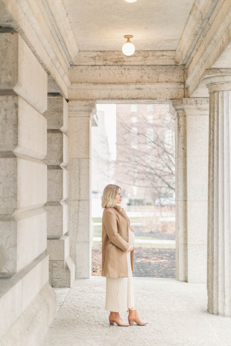Pregnant woman in a long coat and heeled boots stands between tall stone columns during her maternity session in downtown Kansas City.
