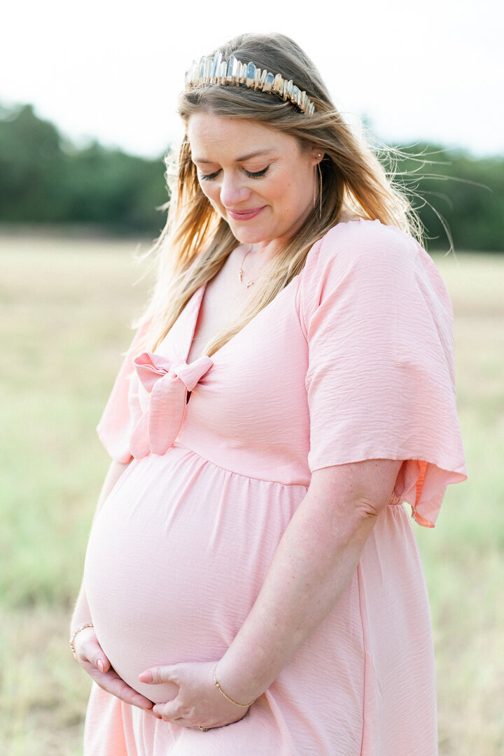 An expectant mother stands in a field at Brushy Creek Lake Park and looks down at her belly.