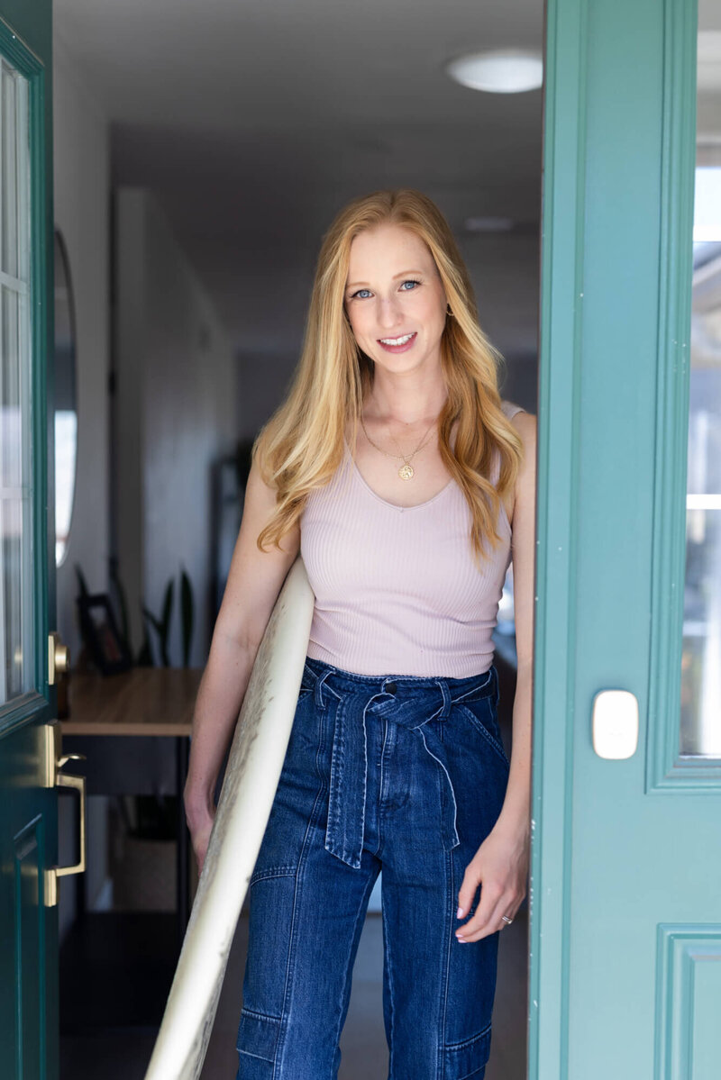 Mary Pernicano, San Diego realtor, standing in her doorway holding a surfboard and welcoming clients into a coastal home — Mary Pernicano Real Estate.