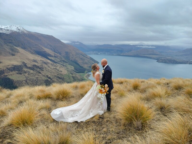 Outdoor wedding ceremony with a couple exchanging vows on a circular platform, surrounded by guests and framed by a stunning lake and mountain backdrop under a partly cloudy sky.