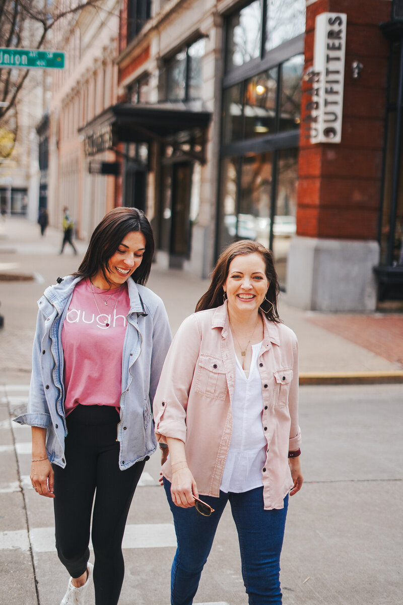 Two women cross the street in a modern city, one wears pink shirt with company logo in white, designed by woman-owned branding company Liberty Type